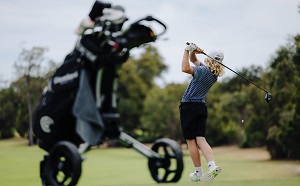 A young person swinging a golf club on a course with a set of golf clubs in the foreground